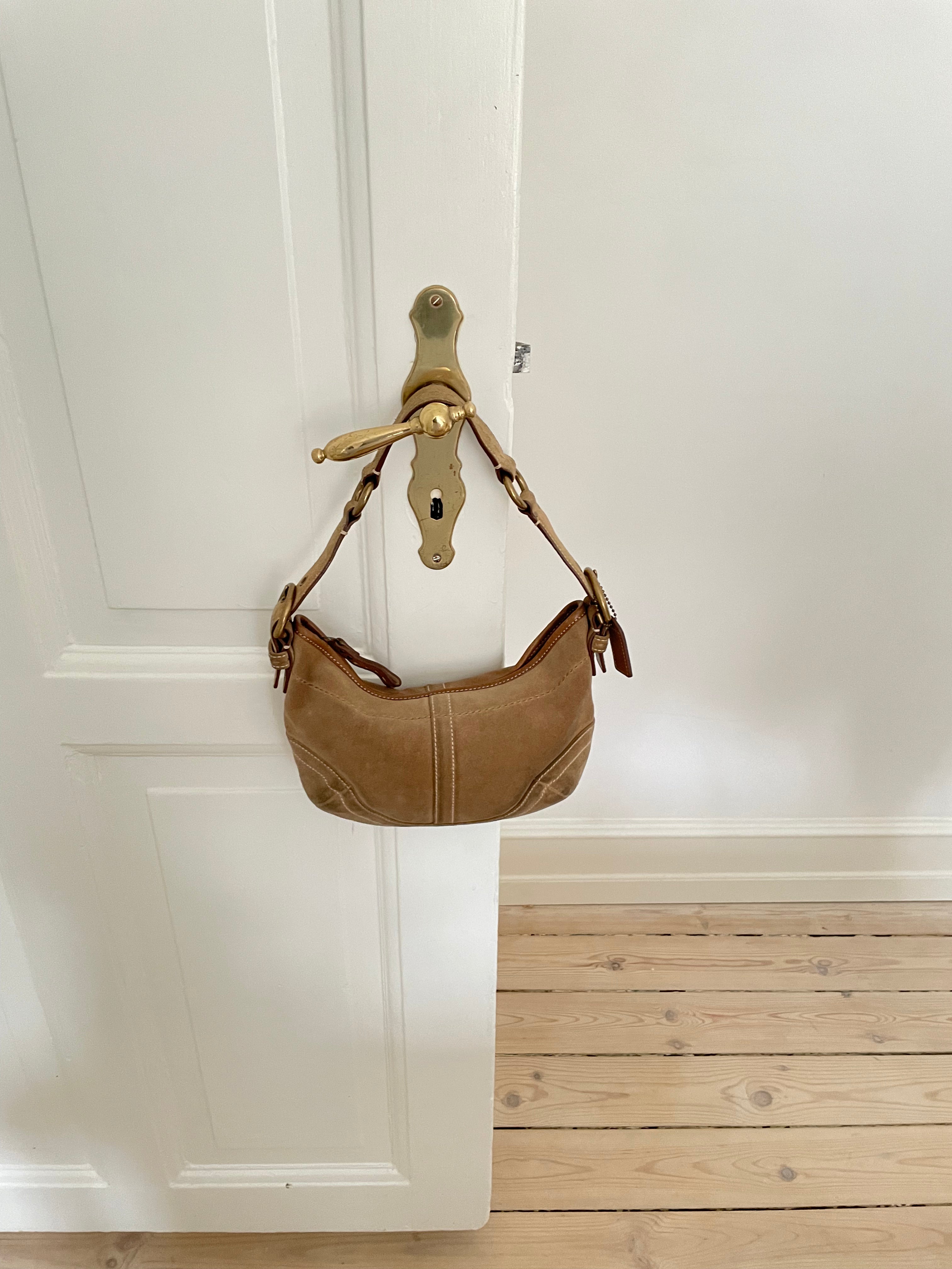 This beautiful Coach shoulder bag, crafted from light brown suede, is in good condition. It features unique white stitching, an adjustable leather strap, and offers an interior zip pocket along with one open pocket.