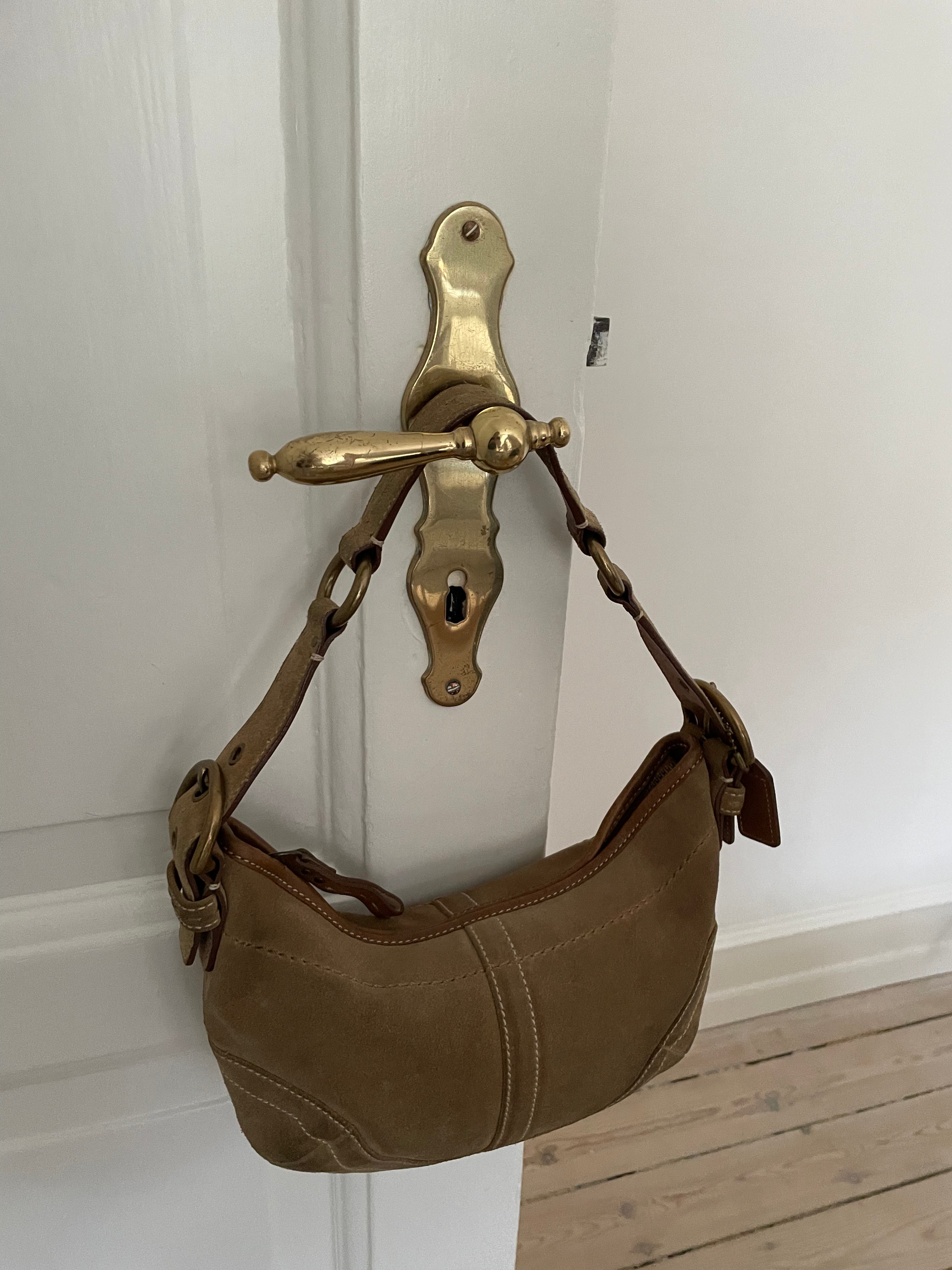 This beautiful Coach shoulder bag, crafted from light brown suede, is in good condition. It features unique white stitching, an adjustable leather strap, and offers an interior zip pocket along with one open pocket.