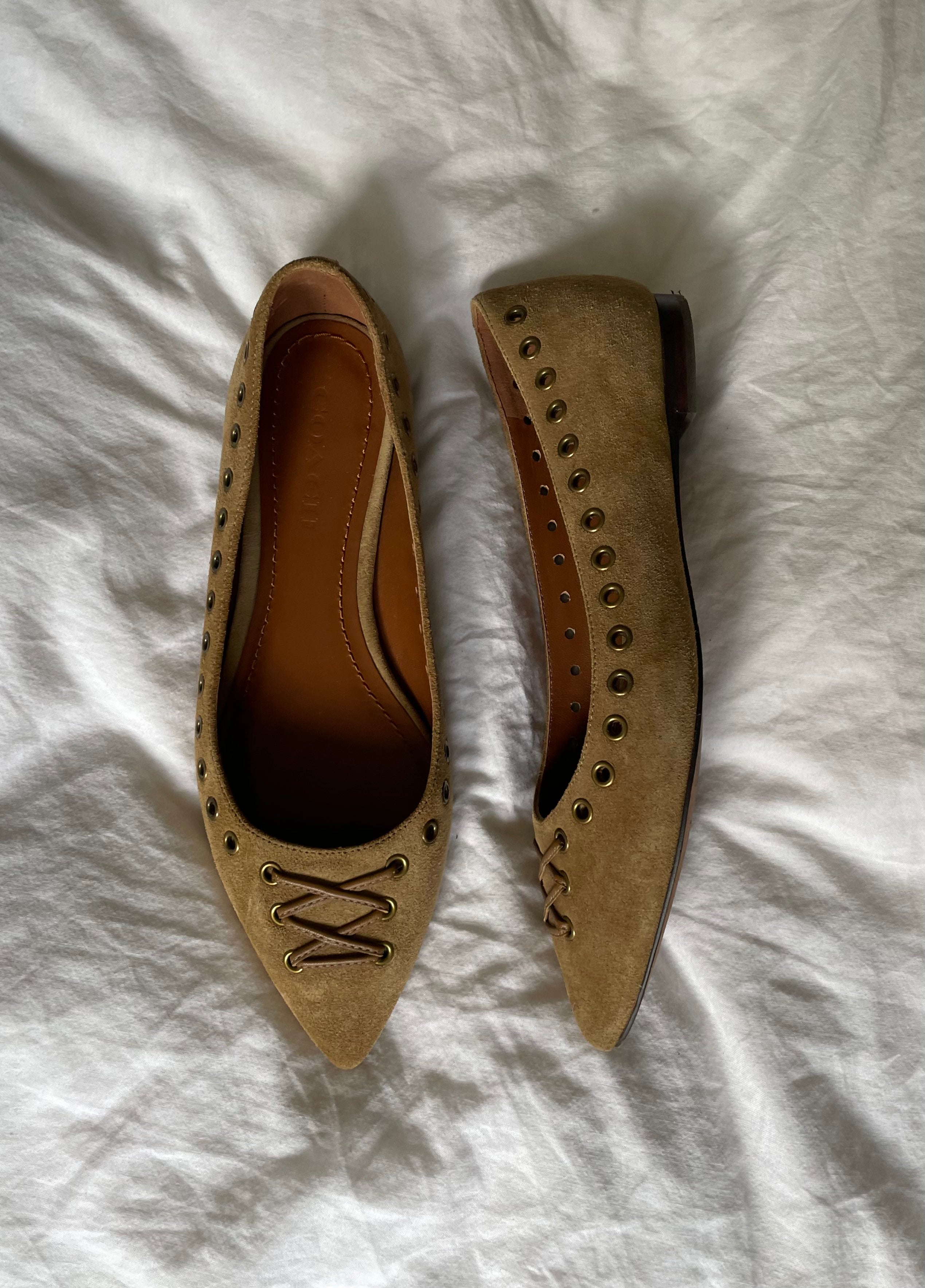 These vintage Coach ballet flats feature a pointed toe, lace-up details, and gold-tone eyelets along the edges. Crafted from beige suede with a smooth leather lining.